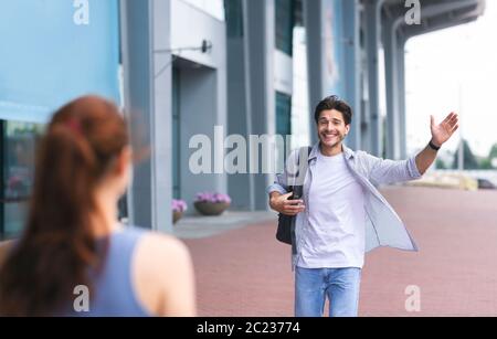 Long-awaited arrival. Happy guy waving hand, greeting girlfriend near airport terminal Stock Photo