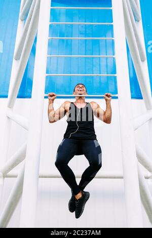 Front view of smiling handsome muscular guy doing pull ups, wearing black sportswear. Sport concept. Stock Photo