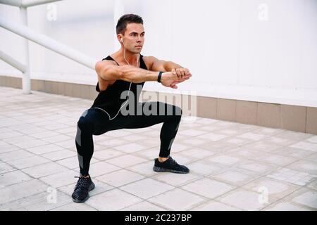 Full length of handsome concentrated sportsman doing squats during workout outdoors, listening to music in earphones. Wearing black sportswear. Stock Photo