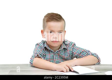 Caucasian school-age boy in a plaid shirt sitting at the table and writing in a piece of paper . there is also a laptop and a phone on the table Stock Photo