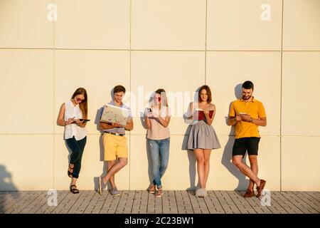 Beautiful young students are using gadgets, reading book and smiling, standing over wall background Stock Photo