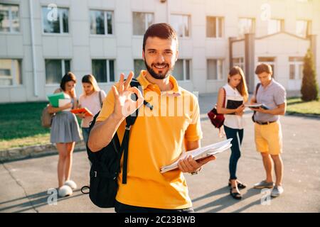 Happy student with note-books and backpack smiling and showing okay while standing on background of university and friends Stock Photo