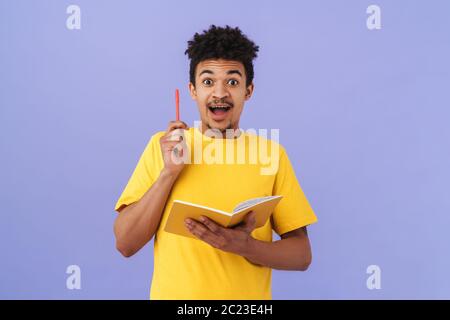 Photo of surprised african american man holding pen and exercise book isolated over purple background Stock Photo