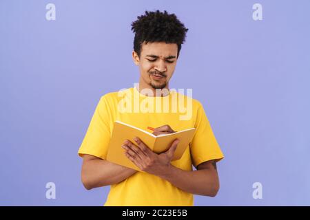 Photo of thinking african american man grimacing and making notes in planner isolated over purple background Stock Photo