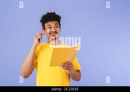 Photo of confused african american man grimacing and making notes in planner isolated over purple background Stock Photo