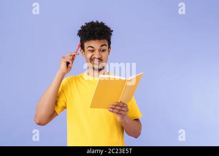 Photo of confused african american man grimacing and making notes in planner isolated over purple background Stock Photo