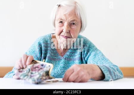 Concerned elderly 96 years old woman sitting at table at home and counting remaining coins from pension in her wallet after paying bills. Unsustainabi Stock Photo
