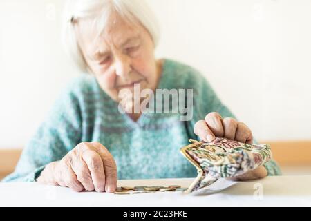 Concerned elderly 96 years old woman sitting at table at home and counting remaining coins from pension in her wallet after paying bills. Unsustainabi Stock Photo