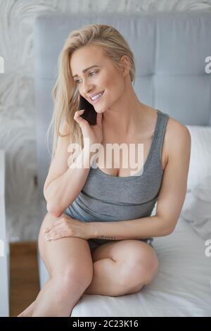 Adorable middle aged blonde woman sitting on bed and talking on mobile phone Stock Photo