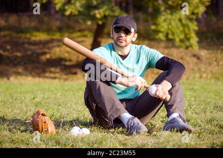 The baseball player is sitting and resting on grass with his sports equipments. Stock Photo