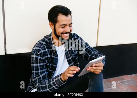 Stylish young hipster man using a digital tablet , cheerfully smiling, looking at tablet screen, sitting on the ground outdoors. Stock Photo