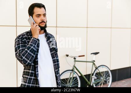 Thoughtful young bearded man communicating by cell phone, while standing outdoors near the bike, wearing stylish shirt. Stock Photo