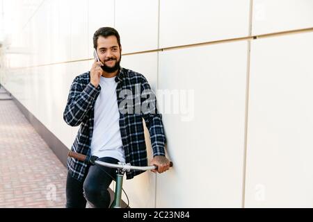 Handsome bearded man having pleasant phone conversation, during sitting on bike, outdoors. Dressed in plaid shirt, t-shirt and jeans. Stock Photo