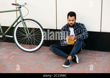 Stylish pensive bearded man sitting with computer tablet near the bike on the ground, reading something, wearing trendy clothes. Outside. Stock Photo