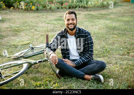 Smiling bearded man resting at park, sitting on grass near the bicycle, looking at camera. Leisure concept. Stock Photo