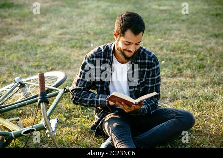 Attractive smiling bearded man reading interesting book, spending time with pleasure while sitting on the grass near the bike. Leisure concept. Stock Photo