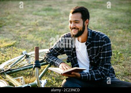Handsome happy bearded man reading interesting book, spending time with pleasure while sitting on the grass near the bike. Stock Photo