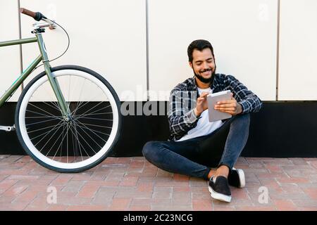 Attractive joyful man using a computer tablet, browsing websites, smiling, while sitting on the ground near the bike. Dressed up in stylish plaid shir Stock Photo