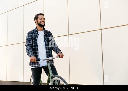 Outdoor photo of cheerful bearded man riding a bicycle , spending time with pleasure, wearing stylish clothes. Stock Photo