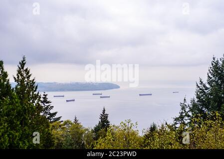 Large cargo ships anchored in Burrard Inlet, just outside the entrance to Vancouver Harbour, viewed from between trees, near Vancouver, BC, Canada. Stock Photo