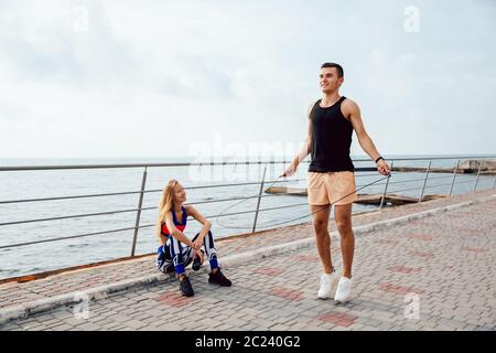 Young sportive couple working out together on the quay, near the sea. Handsome man jumping with skipping rope, while his girlfriend watching at him an Stock Photo