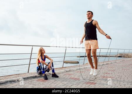 Young sportive couple working out together on the quay, near the sea. Attractive man jumping with skipping rope, while his girlfriend watching at him Stock Photo