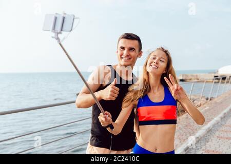Happy attractive couple in sportswear showing a thumb up and peace sign while taking a selfie on mobile phone, after workout outdoors, near the sea. Stock Photo
