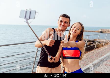 Funny happy couple in sportswear showing a thumb up while taking a selfie on smartphone, after workout outdoors, near the sea. Stock Photo