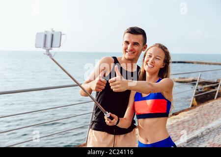 Joyful sportswoman and sportsman in sportswear showing a thumb up while taking a selfie on smartphone, after workout outdoors, near the sea. Stock Photo