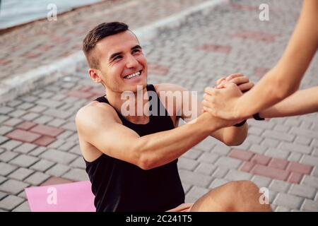 Female helps a handsome sportive man to get up after doing a sport exercise on carpet, support him. Stock Photo