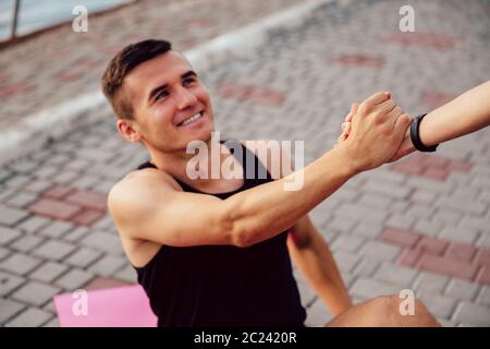 Female helps a smiling man to get up after doing a sport exercise on carpet, support him. Friendship. Stock Photo