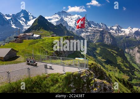 Swiss flag waving and tourists admire the peaks of Monch and Jungfrau mountains on a Mannlichen viewpoint, Bernese Oberland Switzerland Stock Photo