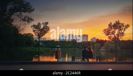 Young couple sitting together on the bench in the park near the lake against bright sunset. Romantic lifestyle background, togetherness concept. Lover Stock Photo