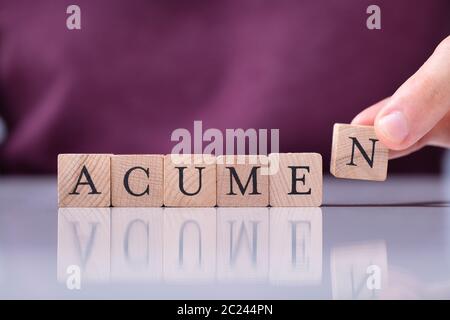 A Person's Hand Placing Last Alphabet Of Word Acumen Over White Reflective Desk Stock Photo