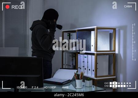 Thief Wearing Balaclava Stealing File From Shelf At Workplace Scene Through CCTV Camera Stock Photo