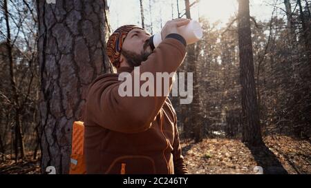 A young tired Caucasian man carries a rest break with a backpack sitting and drinking water on a tree in a nature forest during Stock Photo