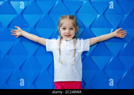 A girl with blond hair in a white T-shirt on a blue background. She smiles and waves her hands Stock Photo