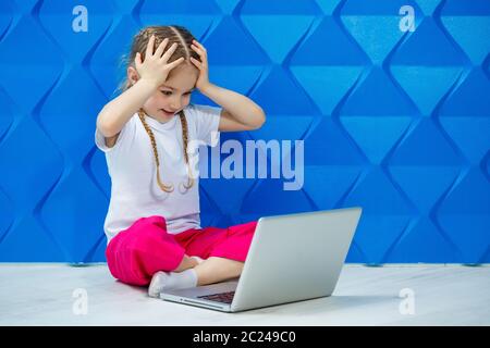 7 years old girl in a white T-shirt sits on the floor with a laptop and presses the keys Stock Photo