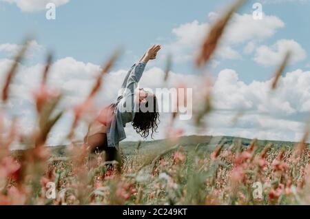 Young woman doing a standing back bend with arms up in flower field framed by flowers leaning in same direction Stock Photo