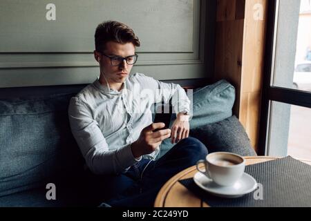 Thoughtful serious man in eyeglasses, sitting on couch in headphones, looking at smartphone screen, spending time at cafe. Stock Photo