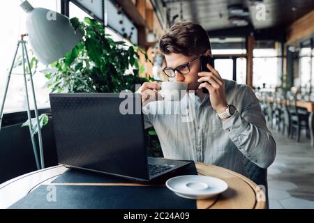 Successful attractive young businessman in eyeglasses drinking coffee, during having phone conversation, looking at laptop screen. At cafe. Stock Photo