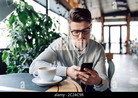 Pensive handsome man in headphones, watching or reading something on mobile phone, drinking coffee, having a break at cafe. Dressed in shirt. Stock Photo
