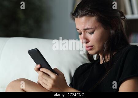 Sad teen crying after read smart phone message sitting on a couch in the dark at home Stock Photo