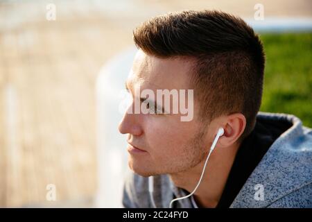 Close-up photo of young pensive man listening to music in headphones, wearing sport jacket, outdoors. Stock Photo
