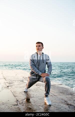 Attractive concentrated guy in sportswear doing exercises for legs, stretching on pier, near the sea. Outdoors. Stock Photo