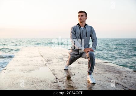 Pensive concentrated man in sportswear doing lunges, stretching, while having workout on pier, near the sea. Outdoors. Stock Photo