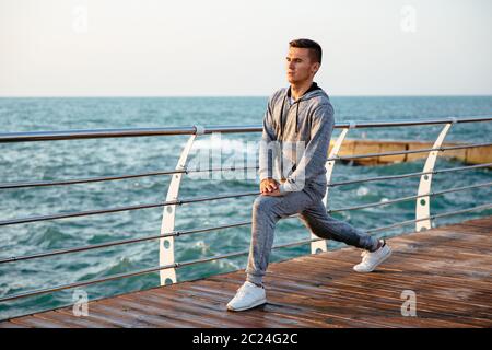 Handsome concentrated man in sportswear doing exercises for legs, stretching on quay, near the sea. Outdoors. Sport concept. Stock Photo