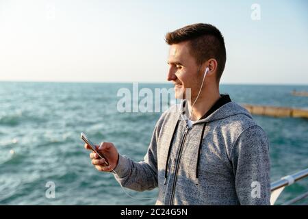 Happy attractive man in sport jacket and headphones, using a smartphone, while walking on quay, near the sea. Stock Photo