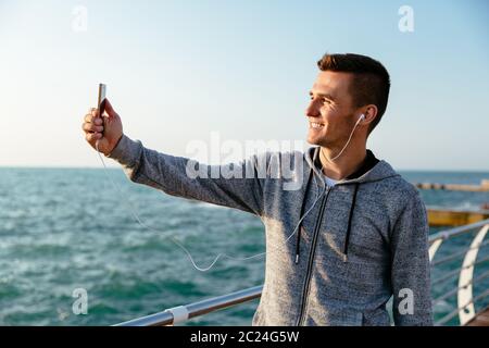 Smiling handsome man in headset and sport jacket takes a selfie on cellphone, while walking on pier. Outdoors. Stock Photo