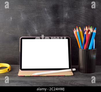 electronic tablet with a blank white screen and multi-colored wooden pencils on a black chalk board background, concept back to school Stock Photo